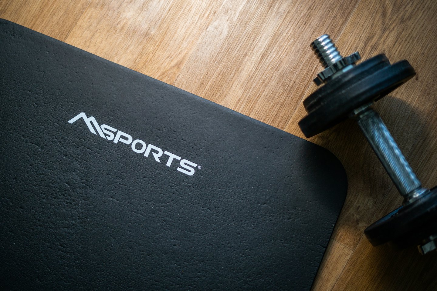 Black exercise mat with dumbbells on a wooden floor.